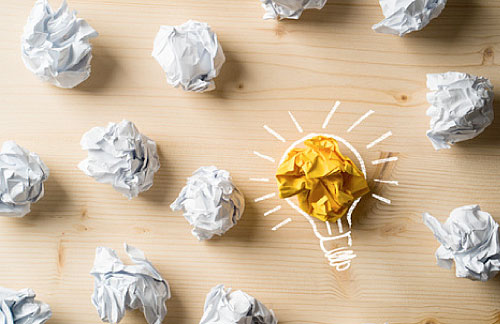 Auf einem Tisch liegen viele zerknüllte Zettel. Darunter einer in Gelb. Jemand hat eine Glühbirne darum gemalt.